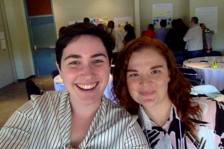 Two women smiling in a selfie, with a group of people standing behind them working collaboratively at posters on a wall.